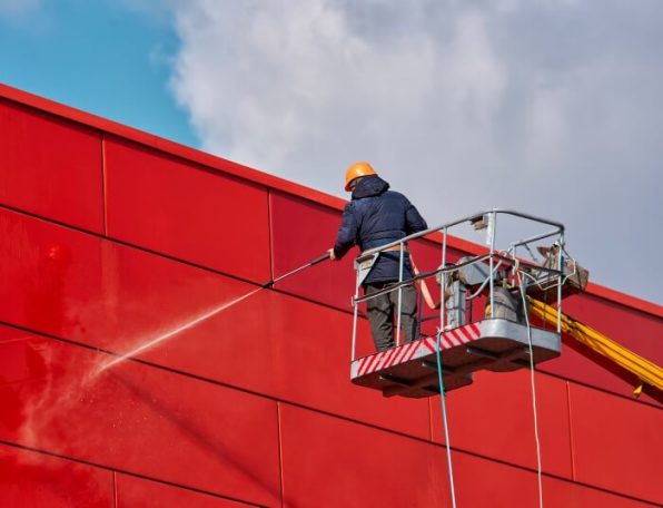 Commercial Cleaning A worker in a lift cleaning a red wall with a pressure washer against a cloudy sky.